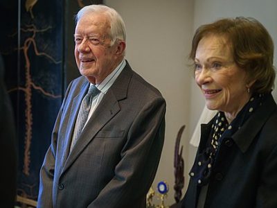 An elderly Jimmy and Rosalynn Carterare shown smiling, indoors - Jimmy wears a dark suit and blue tie while Rosalynn wears a black jacket with a polka dot blouse