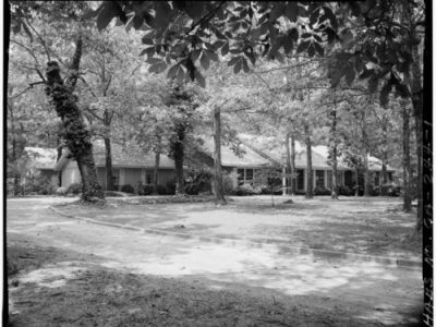 Black and white photograph of the Carter’s ranch-style family house in Plains, Georgia with a dirt driveway, surrounded by tall trees casting dappled shadows