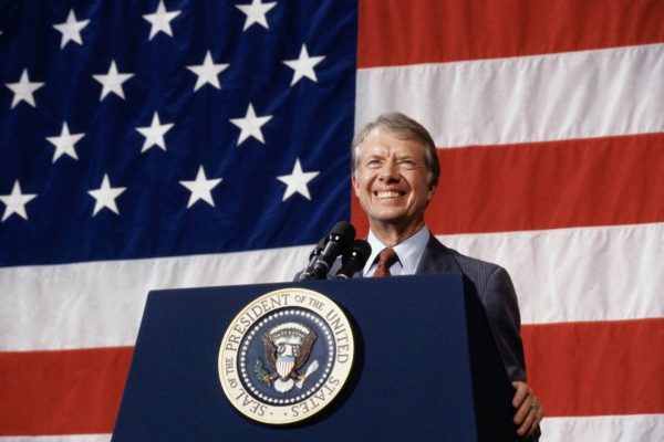 A broadly smiling President Jimmy Carter person stands behind a dark blue podium bearing the Seal of the President of the United States. He is speaking into multiple microphones, with a large American flag serving as the backdrop, suggesting the formal and significant nature of the setting, such as an official speech or announcement