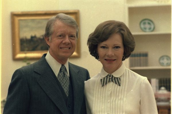 A warm portrait photograph from the 1970s of Jimmy and Rosalynn Carter during his presidency. The couple, known for their 77-year marriage and lifelong dedication to service, family values, and caregiving, stand together against a light-colored wall with shelves. President Carter wears a dark suit with a tie, and First Lady Rosalynn Carter wears a white blouse with a striped bow. Both are smiling warmly at the camera, embodying the genuine warmth and compassion that characterized their work in mental health advocacy, humanitarian causes, and their commitment to home and family. A framed artwork and books are visible on shelves in the background.