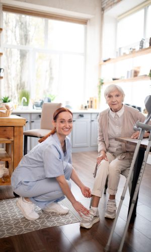 Smiling female in-home caregiver with medium-length brunette hair pulled back into a ponytail, dressed in light-blue scrubs and white trainers crouching down next to an elderly woman to assist her with her shoes. The older woman has white hair cut in a short Pixie style, and is dressed in a pale beige cardigan and top, cream-color pants, and pastel brown and white trainers. The older woman is seated in a chair next to a table in her kitchen at home, with a mobility aid visible nearby