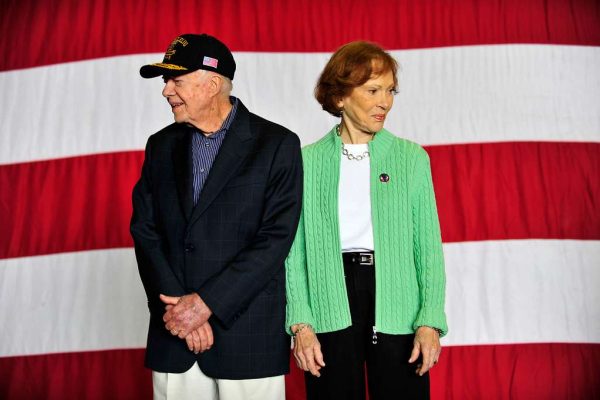 Jimmy and Rosalynn Carter in their later years standing in front of an American flag backdrop, Jimmy wearing a navy blazer and baseball cap, Rosalynn in a green cardigan, white top, black pants and a belt