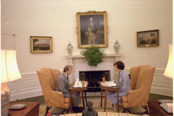 Jimmy and Rosalynn Carter sitting in wingback chairs at a small dining table in front of a fireplace in an elegant White House room, with framed portraits and art visible on the walls