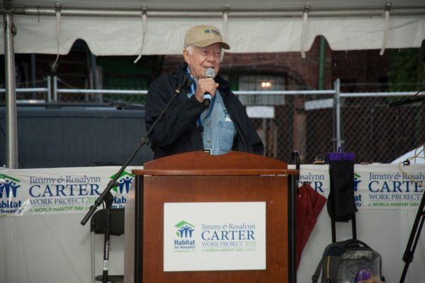 A smiling Jimmy Carter wearing a Habitat for Humanity cap speaks with a microphone at a podium during a Carter Work Project event